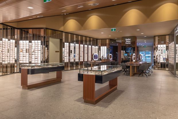 A refined commercial showroom interior with illuminated wall displays, freestanding glass-topped timber-and-dark-finish display counters, consultation tables, and a sculptural ribbed timber ceiling feature. The space has polished stone or terrazzo-style flooring, warm integrated lighting, and extensive glazed or reflective display partitions.