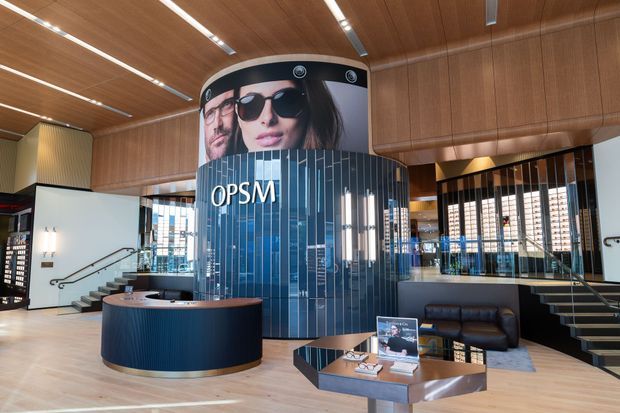 A polished commercial optical showroom and reception area featuring a curved blue reception desk, a cylindrical blue display tower with large OPSM branding and eyewear advertising, dark vertical feature tiles, timber wall and ceiling panelling, glass partitions, integrated linear lighting, and a small lounge seating area.