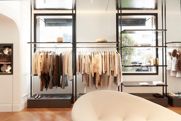 Bright, refined walk-in wardrobe or boutique-style dressing room with open black metal clothing racks, neatly arranged neutral-toned garments, integrated low storage, shelving, large black-framed windows, and warm timber flooring. A curved upholstered seating piece occupies the foreground, while a white arched opening and display shelving add architectural detail.