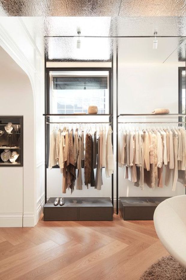 Bright, refined walk-in wardrobe with floor-to-ceiling mirrored and glazed wardrobe joinery, hanging rails filled with clothing, integrated low drawers, display shelving, and warm herringbone timber flooring. A curved upholstered furniture edge is visible in the foreground.
