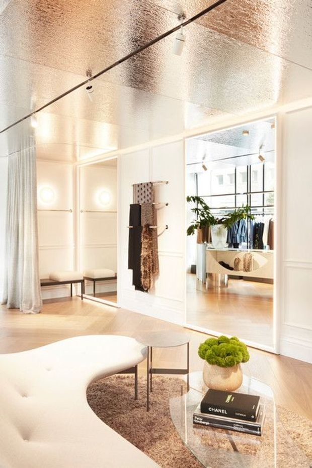 Bright, refined retail interior with a white paneled fit-out, reflective metallic ceiling, pale timber flooring, large mirrors, and sculptural cream seating. Clothing displays and hanging garments are visible in the adjoining mirrored retail area. The foreground includes a curved sofa, small round tables, a glass coffee table, and soft neutral furnishings, with track lighting and decorative wall lighting.