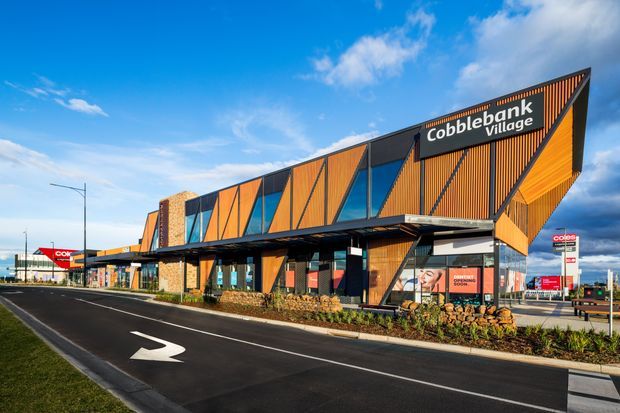 Exterior street view of Cobblebank Village, a contemporary commercial shopping-centre building with a dramatic angular facade. The building features dark metal framing, warm orange timber-look vertical cladding, stone masonry accents, extensive glazing, projecting roof forms, and prominent retail signage. A paved roadway and landscaped frontage are visible in the foreground.