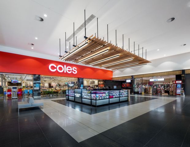 Bright contemporary retail shopping-centre interior featuring a prominent Coles supermarket frontage, deli and food displays, refrigerated promotional island units, polished tiled flooring, and a large suspended timber slat feature ceiling with integrated lighting.