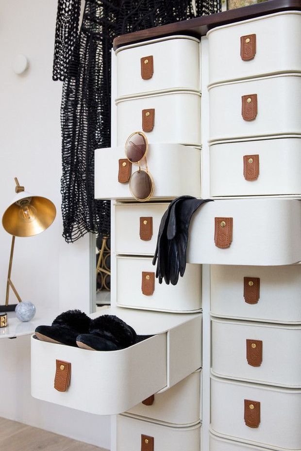 Close-up interior view of a tall modular drawer storage system with rounded white drawer fronts, dark timber-coloured tops, and brown leather pull tabs. One drawer is open with black footwear inside, while black gloves and sunglasses rest on the unit. A black woven pendant or decorative screen, brass floor lamp, mirror, and pale timber flooring add a stylish dressing or wardrobe atmosphere.