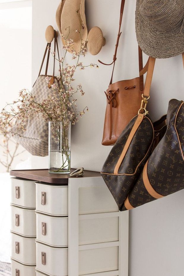 A styled entryway vignette with a cream-coloured drawer cabinet beneath a dark timber top, displaying a glass vase with delicate branches. Handbags and woven accessories hang on wall-mounted hooks and pegs above and beside the cabinet. The image has a warm, curated, fashion-oriented aesthetic.