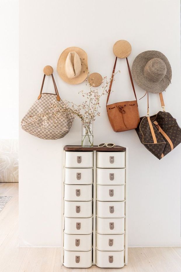 Bright, styled entryway vignette with a tall white drawer cabinet against a white wall, decorated with woven hats and hanging leather and patterned bags. A glass vase with delicate branches and sunglasses sit on the cabinet, while pale timber flooring and a partial adjacent room are visible.