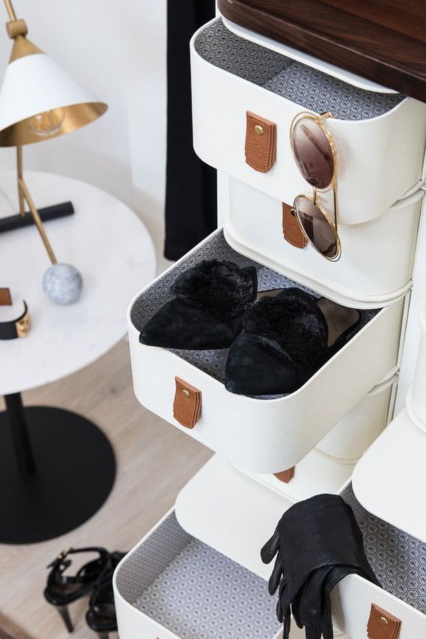 Close-up interior view of a stylish dressing or bedroom corner featuring stacked white storage trunks with patterned lining and leather pulls, displaying black accessories including gloves, slippers, and sunglasses. A white round side table with a modern gold-toned lamp and black accents is visible in the background.
