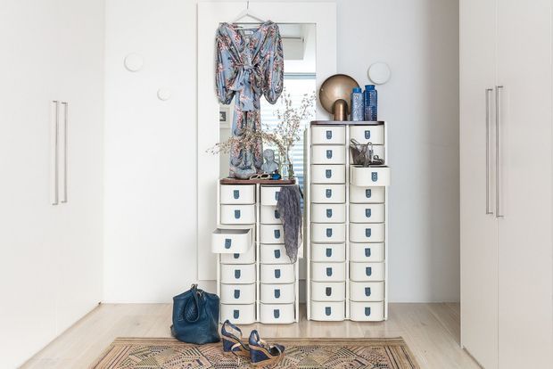 Bright, minimalist walk-in wardrobe or dressing area with extensive white modular drawer cabinetry, tall built-in cupboards, long vertical handles, and a central full-length mirror. Clothing, shoes, handbags, and decorative accessories are arranged around the storage units. Pale timber flooring and a patterned rug add warmth to the otherwise white space.
