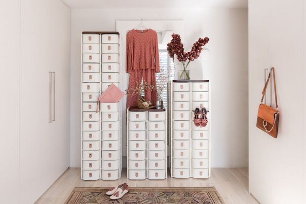 A bright, compact walk-in wardrobe or dressing room with extensive white drawer cabinetry, hanging garments, handbags, shoes, and a patterned floor rug. The room has white walls and built-in wardrobes, with a light timber-look floor and soft pink and terracotta clothing and accessories.