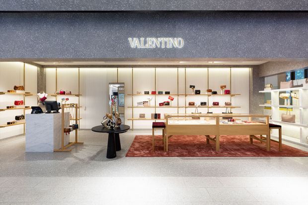 A refined Valentino retail boutique interior viewed from the mall corridor. The space features illuminated vertical wall panels, pale display shelving, glass-topped and timber-framed product display tables, a marble-look checkout counter, and a dark textured fascia with illuminated branding. The palette is predominantly white, cream, light wood, and warm gold, with a large muted red patterned rug anchoring the central display.