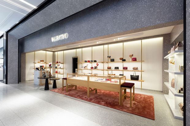 A refined luxury retail boutique interior with a warm, illuminated display wall, open shelving, glass-topped timber display tables, and a deep red area rug. The storefront is framed by dark speckled wall surfaces and a black fascia bearing a brand logo, creating a high-contrast, gallery-like commercial setting.