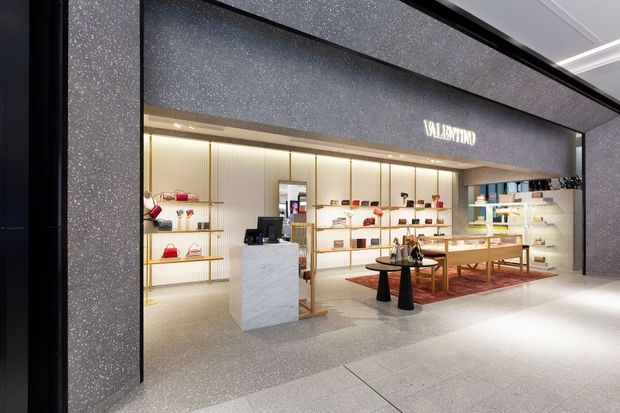 A refined luxury retail boutique storefront in a shopping centre, featuring a dark speckled stone-look portal, bright illuminated display shelving, a marble-look reception counter, timber display tables, and carefully arranged fashion accessories. The Valentino sign is mounted above the open shopfront. The space has a polished, contemporary high-end commercial fit-out.