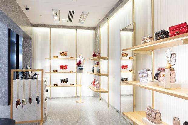 Bright, refined retail boutique interior displaying handbags and small leather goods on open shelving, wall-mounted shelves, and a central display rack. The space features white vertically ribbed wall panels, light timber shelving with slim brass-toned supports, reflective surfaces, a terrazzo-like floor, and integrated ceiling lighting.
