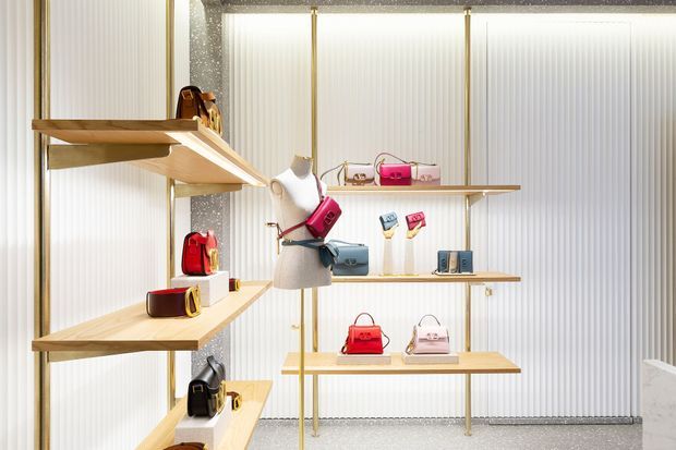 A bright, refined retail display interior featuring tiered light-wood shelving, gold-toned supports, white ribbed translucent wall panels, and a central mannequin displaying handbags. Multiple colourful handbags and accessories are arranged on the open shelves, with a polished terrazzo-like floor and ceiling edge visible.