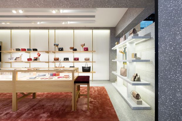 A refined luxury retail showroom displaying handbags, accessories and small leather goods. The space features illuminated wall shelving, glass-topped timber display cases, pale vertical wall panelling with brass-toned trims, terrazzo-like grey surfaces, recessed linear lighting and a deep red area rug.