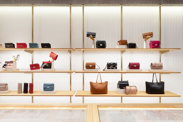 A brightly lit retail display wall showcasing handbags and small leather goods arranged on multiple pale timber shelves. The shelving is framed with slim brass-toned vertical supports against a ribbed white backdrop, with illuminated glass display counters along the bottom.