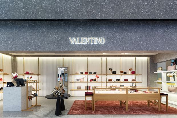 Interior view of a Valentino luxury retail boutique, featuring illuminated vertically ribbed display walls, pale stone checkout or display counters, timber-and-glass display tables, open shelving with handbags and accessories, and a deep red patterned area rug. The space has a refined, high-end showroom atmosphere with dark textured ceiling and fascia surfaces.