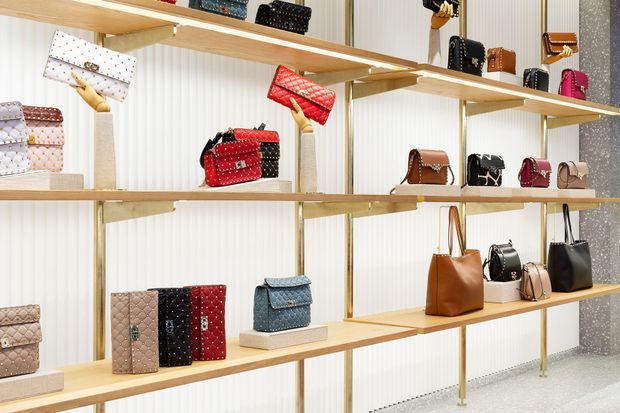 A refined retail display wall showcasing handbags and small leather goods arranged on light timber shelves with slender brass-toned supports. The backdrop features vertical ribbed white paneling, with polished light flooring and a glittering grey feature wall at the side. The composition is bright, orderly, and luxurious.