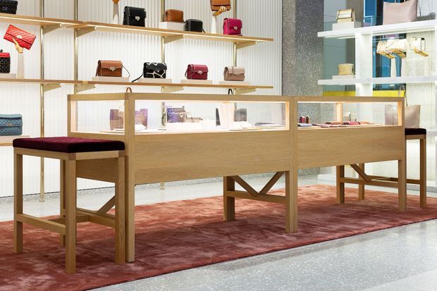 A refined luxury retail interior displaying handbags and accessories on open wall shelving and illuminated glass-topped timber display counters. The space features pale timber joinery, white ribbed wall panels, a dark textured feature wall, terrazzo flooring, and a muted red patterned area rug with matching burgundy seating.