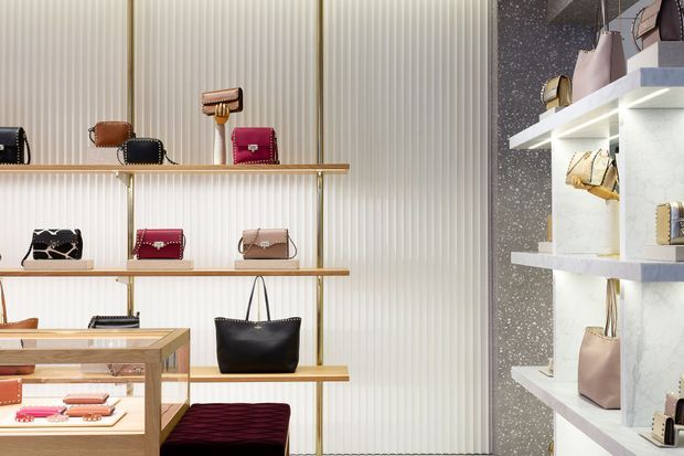 Luxury handbag and accessories retail display featuring handbags arranged on slim brass-trimmed wall shelves, a glass-topped wooden display counter, and illuminated white shelving against a vertically ribbed wall and dark speckled feature surface.