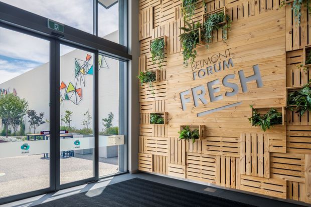A bright commercial entry foyer featuring a large black-framed glazed door and floor-to-ceiling windows beside a prominent timber slat feature wall. The wall incorporates layered crate-like timber panels, integrated planter boxes with trailing greenery, and raised signage reading “Belmont Forum FRESH.” Outdoor seating and geometric artwork are visible through the glazing. The space has a contemporary, natural-material retail or shopping-centre character.