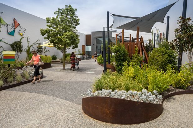 A landscaped outdoor courtyard or public garden space with curved rusted metal planter beds, gravel-textured paving, mature and ornamental planting, colourful abstract wall art, and sculptural shade sails supported by dark posts. A timber play or seating structure and modern building walls are visible in the background. The scene is bright, contemporary, and designed for pedestrian use.