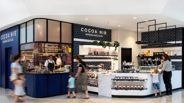 Interior view of a contemporary artisan chocolate shop or café retail space. The fit-out features a dark navy and black glazed service counter on the left, black subway-tile walls, white open shelving, a pale marble-look central counter, integrated product displays, and warm illuminated cabinetry. Customers and staff are present, with packaged chocolate products displayed throughout.