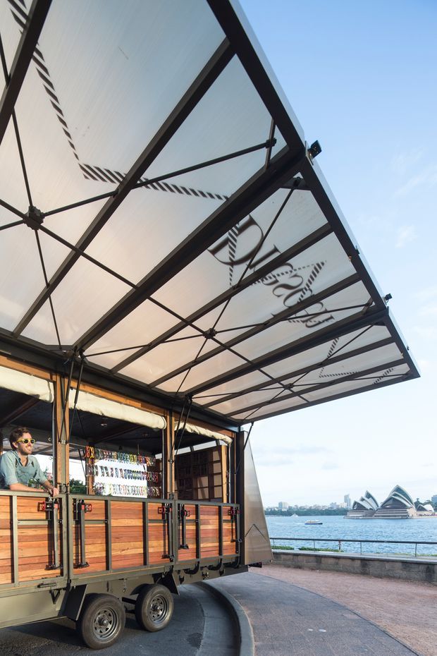 Exterior view of a mobile food or beverage trailer with a large retractable canopy, timber-clad service counter, black metal framing, and displayed bottles. It is parked on a waterfront promenade with a visible harbour and opera-house-like landmark in the background.