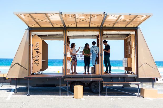 A compact modular exhibition or retail space mounted on a trailer, photographed open on a beachfront promenade. The structure has folding timber-framed wall panels and a raised hinged roof canopy, creating an open-air interior where several people are gathered. Built-in shelving and display elements are visible inside, with a bright ocean and beach setting behind it.