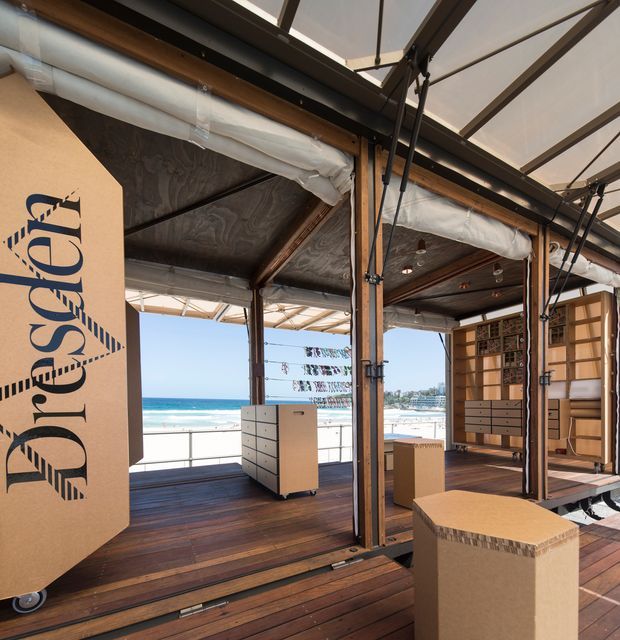 A bright open-air commercial display or event space on a timber deck beside the beach. The structure features exposed timber posts and beams, dark timber decking, translucent retractable roof or awning panels, visible services, and movable cardboard display units. Ocean, sandy beach, and waterfront buildings are visible beyond the open facade.