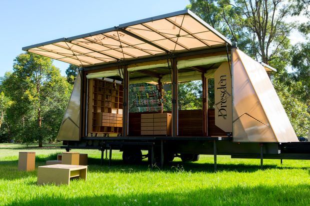 A compact, elevated mobile architectural structure or exhibition pavilion set in a grassy park-like landscape. The open-sided timber-framed unit has translucent fabric or polycarbonate canopy and angled side panels, with built-in timber shelving, drawers, and display surfaces inside. It sits on a dark trailer-like base with visible wheels, and simple timber box seating is arranged on the lawn in front. Bright daylight and surrounding trees create an outdoor, temporary pavilion setting.