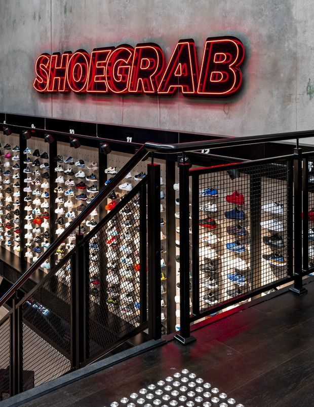 Interior view of a multi-level sneaker retail store, featuring extensive wall-mounted shoe displays, a large red neon-style “SHOERAB” sign, black metal mesh stair and mezzanine railings, concrete-look walls, track lighting, and dark wood flooring.