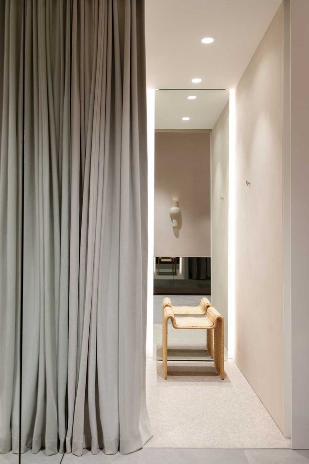 A softly lit minimalist interior corridor or dressing area with floor-to-ceiling pale grey curtains on the left, smooth warm-white walls, recessed ceiling lights, and a pale carpeted floor. A sculptural woven timber chair sits in the passage, with a wall-mounted decorative object and a reflective surface visible at the far end.