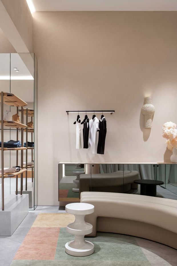 A refined contemporary fashion retail interior with a neutral plastered wall, mirrored shelving and display cabinetry, a minimalist clothing rail, sculptural wall-mounted bust, curved upholstered seating, and a small white pedestal table. Soft integrated lighting and muted pastel rug panels create a calm, luxurious atmosphere.