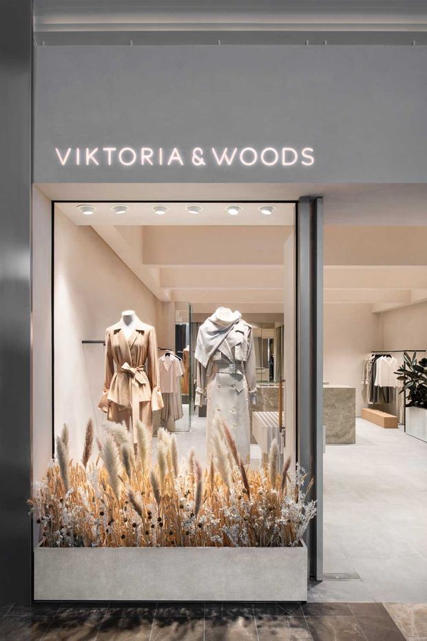 A contemporary fashion retail storefront for Viktoria & Woods, featuring a large glazed entrance, illuminated signage, mannequins in neutral clothing, pale interior finishes, integrated ceiling spotlights, and a prominent concrete planter filled with dried ornamental grasses at the threshold.