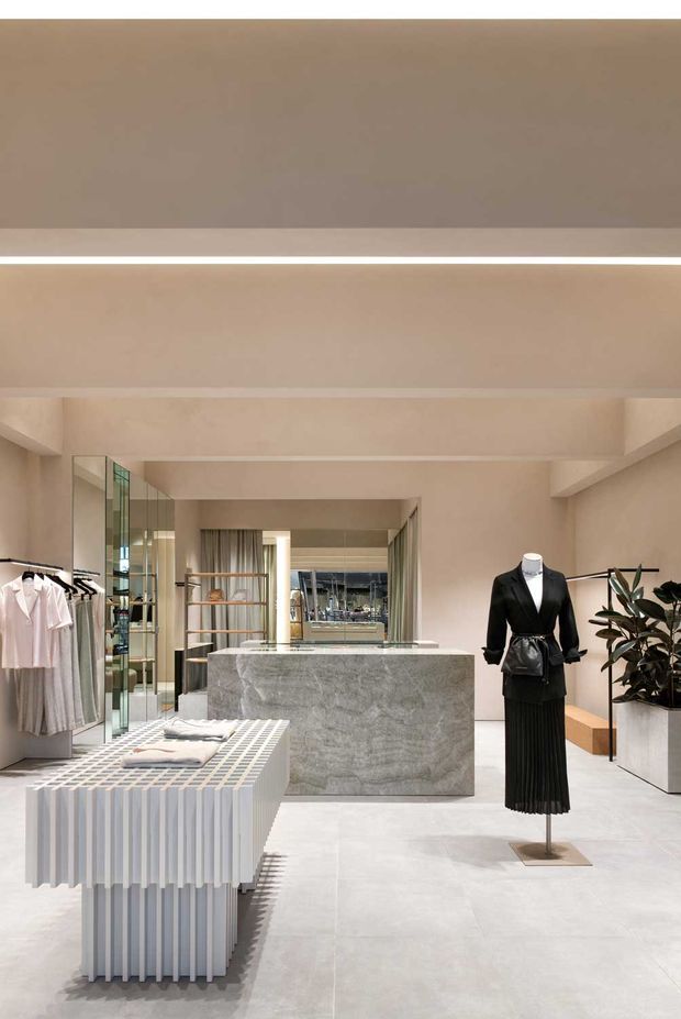 A refined contemporary fashion retail interior with a softly monochromatic beige and grey palette. The space features a sculptural grey stone-clad central counter, pale tiled flooring, mirrored and glazed display elements, minimalist clothing racks, a pleated display plinth, recessed linear lighting, and a mannequin dressed in black. Built-in shelving and greenery add visual depth while the open layout creates a calm, gallery-like atmosphere.