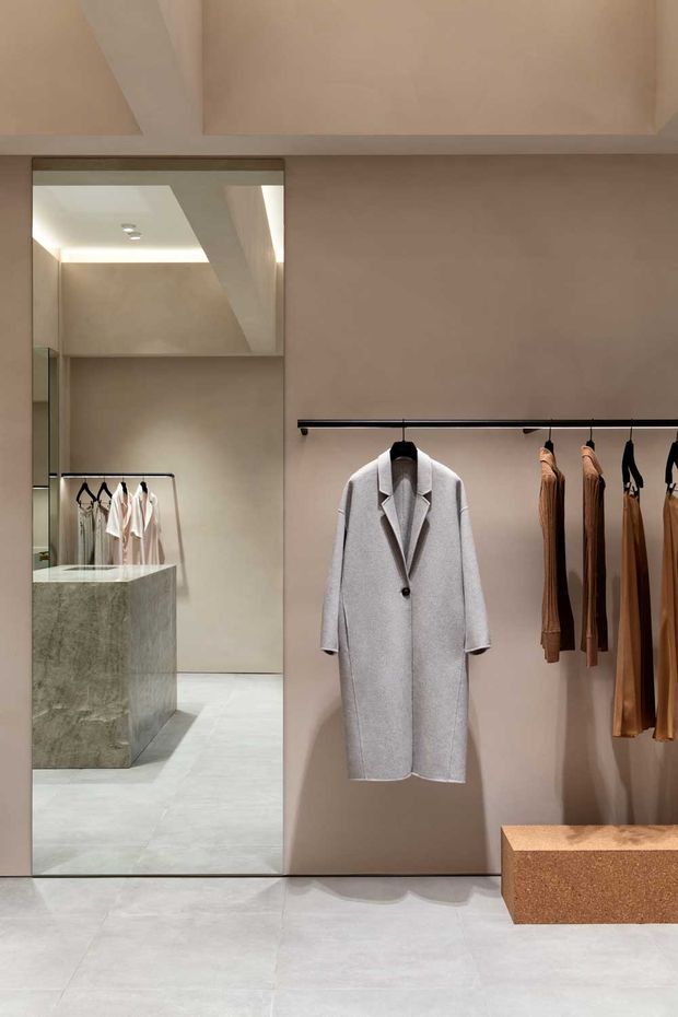 A refined minimalist clothing boutique or walk-in wardrobe with warm taupe plaster walls, pale stone-look tiled flooring, a full-height mirror, a sculptural stone display island, and black rail-mounted clothing racks. Neutral garments and a long light-grey coat are displayed alongside a low bronze-toned bench or plinth. Integrated lighting and clean geometry create a calm, gallery-like retail interior.