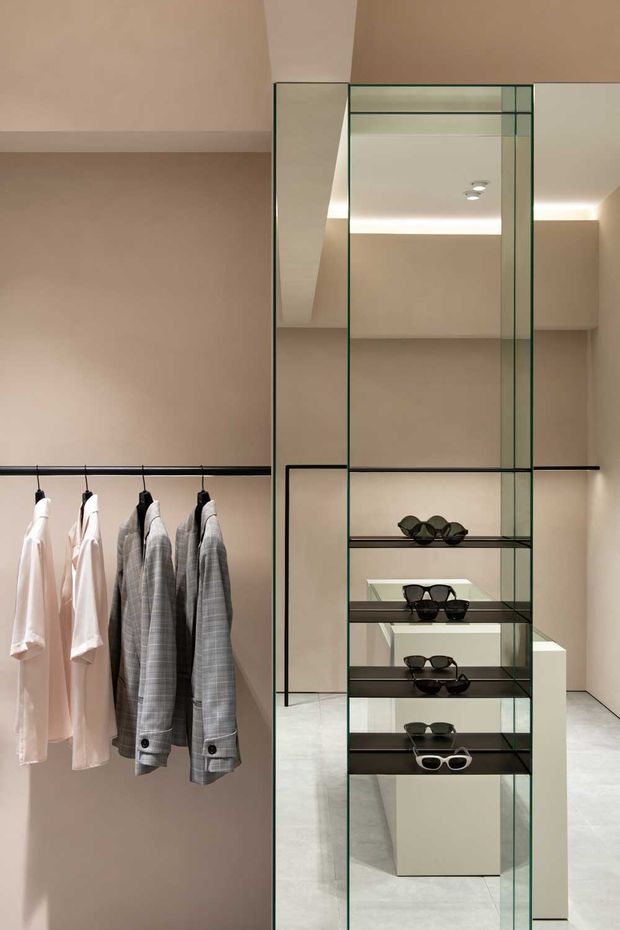 A refined walk-in wardrobe or boutique dressing room with floor-to-ceiling framed glass cabinetry and open shelving displaying sunglasses. Black hanging rails hold light shirts and grey checked jackets against warm beige walls. Integrated linear and recessed lighting, pale flooring, and a central white display island create a minimalist luxury retail-like setting.