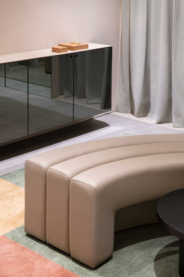 A refined contemporary living room vignette featuring a curved beige upholstered sofa in the foreground, a dark mirrored sideboard with a pale benchtop along the wall, floor-length sheer curtains, and a muted geometric area rug. The palette is soft and neutral with dark reflective cabinetry and rounded furniture forms.