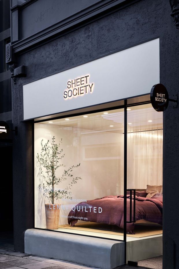 Street-level storefront for “Sheet Society,” featuring a dark textured facade, illuminated white projecting signage, large glass display window, and a warmly lit bedroom display with a bed dressed in muted mauve bedding and a potted olive tree.