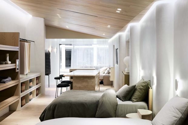 A bright, compact open-plan studio-style bedroom with a bed in the foreground, built-in timber shelving and wardrobe storage along the left wall, a small seating area, and a compact kitchenette or work surface toward the rear. The room features white sculpted wall niches, warm timber ceiling lining and flooring, recessed lighting, and soft neutral bedding.