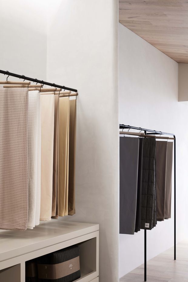 A minimalist textile showroom displaying neutral-toned fabric samples on black metal garment rails. Built-in white cabinetry with organized storage boxes occupies the foreground, while pale timber flooring and ceiling add warmth to the bright, pared-back commercial interior.