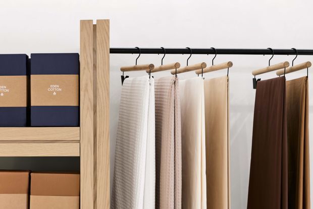 A minimalist textile retail display featuring neutral-toned fabric samples hanging from a black metal rail, with light timber vertical supports and shelving containing neatly packaged textile products. The composition has a clean, warm, contemporary showroom aesthetic.