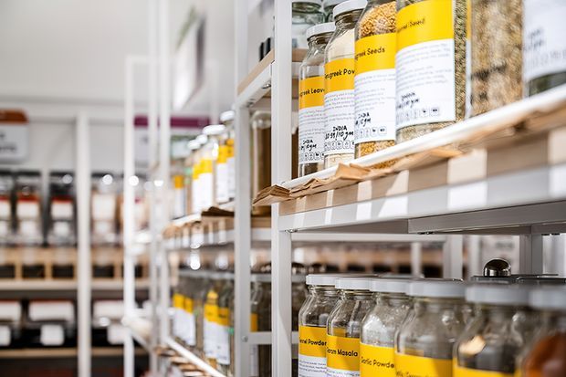 Close-up view along tall open shelving in a pantry or bulk-food storage area, with rows of labelled glass jars and containers holding dry goods. The shelving has light-coloured timber shelves and white structural supports, creating a bright, organised storage environment.