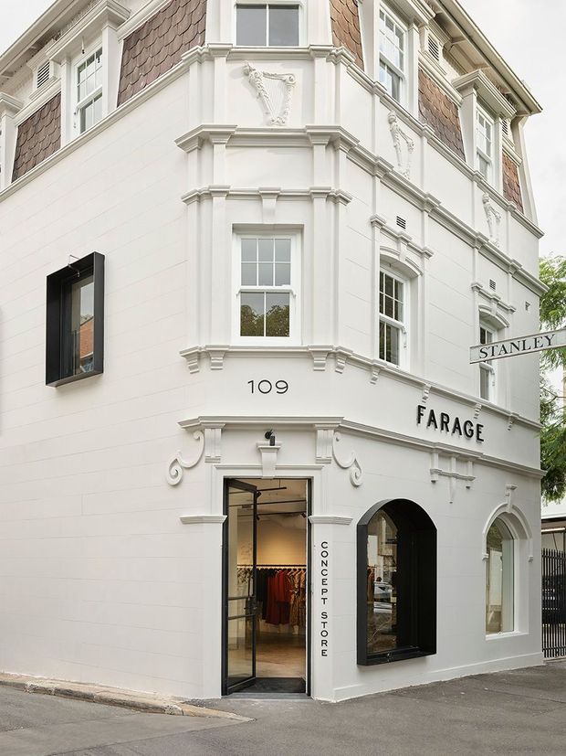 Street-level exterior view of a refined multi-storey corner commercial building with a white rendered facade, ornate classical mouldings, bay windows, arched openings, decorative motifs, and upper sections clad in reddish-brown shingle-like material. Signage identifies the space as a concept store and garage, with clothing visible through the entrance.