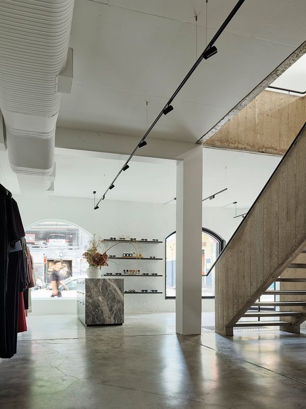 A bright, minimalist retail interior with polished concrete flooring, exposed concrete stair construction, white structural columns, suspended black track lighting, and a large exposed ventilation duct. Clothing racks and display shelving occupy the open showroom, with a stone-clad display island near the centre and arched windows bringing in natural light.