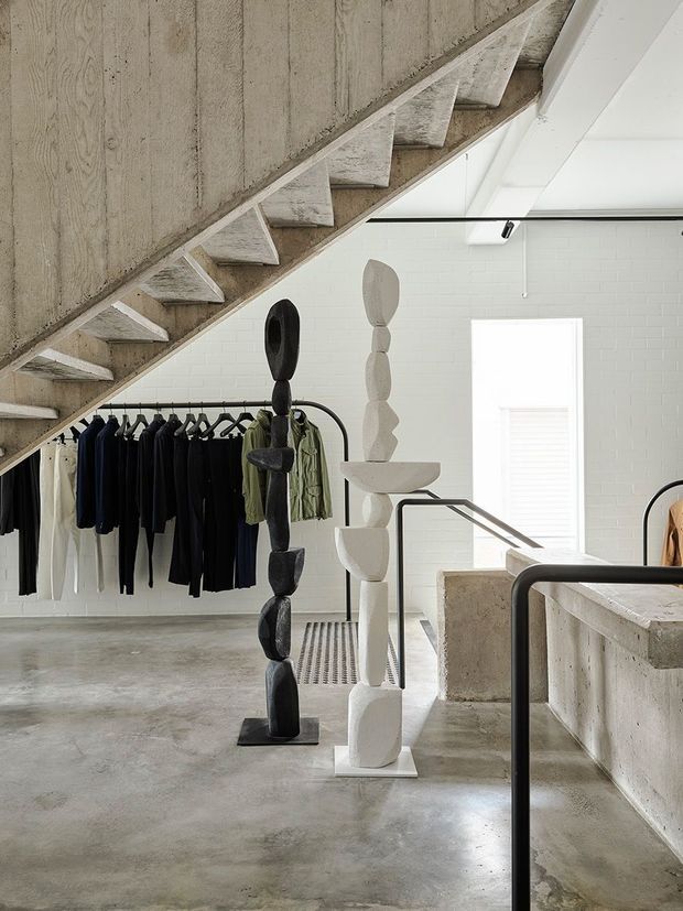 A minimalist industrial retail interior beneath a concrete stairway, with exposed raw concrete surfaces, white painted brick walls, black metal railings, clothing racks, and two sculptural display figures in contrasting black and white. The space has polished concrete flooring and a bright, restrained monochromatic palette.
