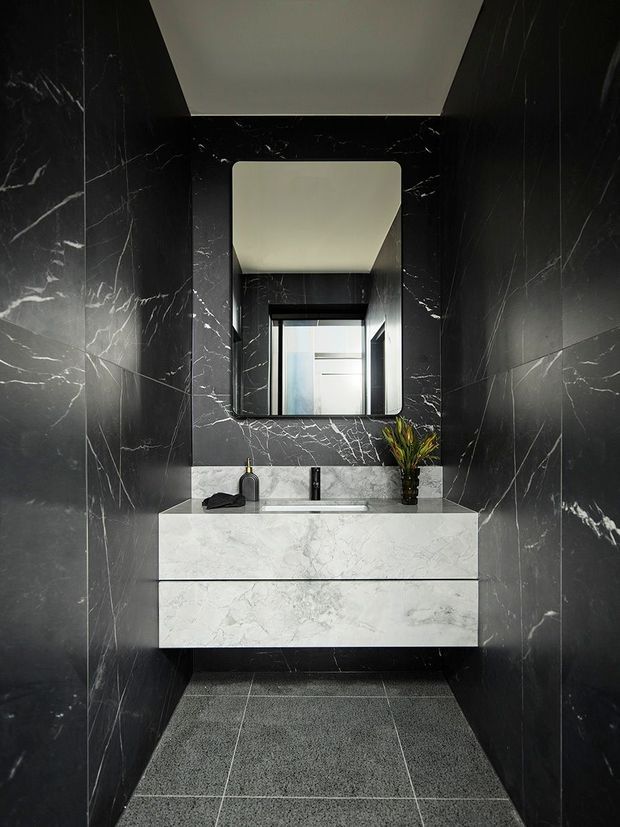A compact, dramatically styled bathroom or powder room with floor-to-ceiling dark marble-look wall slabs, a floating white marble vanity with integrated basin, black tapware, a rounded-rectangle mirror, and dark textured floor tiles. A narrow window brings in daylight behind the mirror, while a small floral arrangement adds a natural accent.