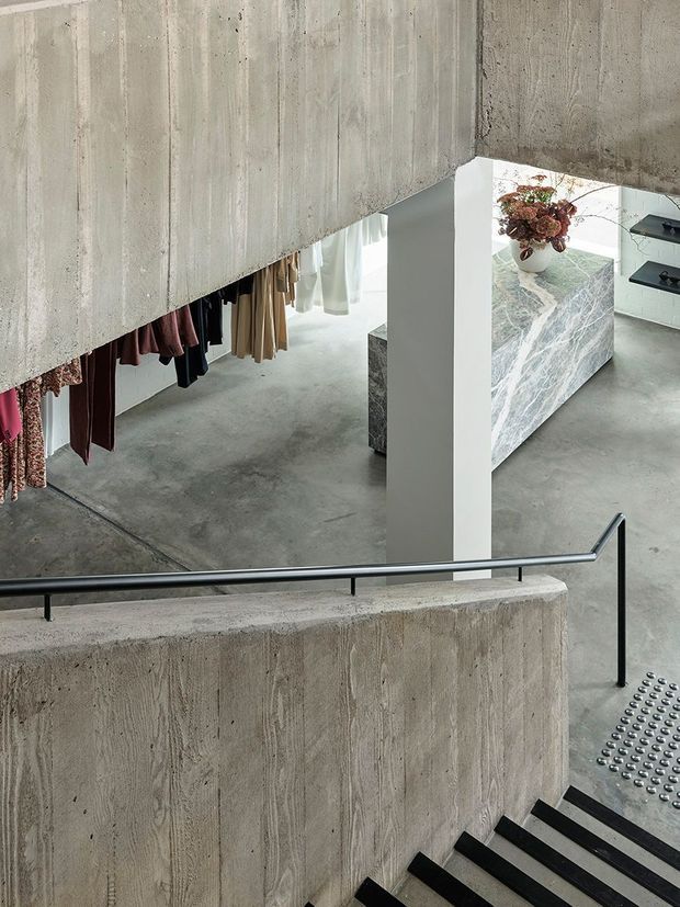 A sculptural interior stairway descends through a raw concrete space into an open-plan kitchen and dressing area. The scene features board-marked concrete walls, floors and stair balustrades, a slim black metal handrail, black stair treads, hanging garments, and a dramatic veined stone kitchen island. A tactile metal floor strip provides stair-edge guidance, while daylight enters through sheer curtains.