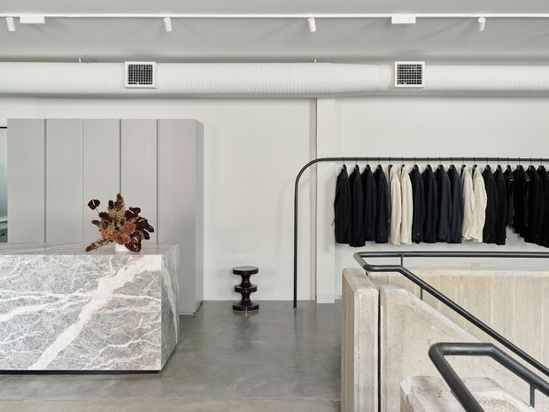 A minimalist contemporary clothing retail interior with a polished grey concrete floor, pale built-in cabinetry, a dramatic veined stone display counter, exposed white ductwork, and black metal clothing rails. Concrete stair or display plinth elements with dark handrails occupy the foreground, while a restrained monochrome garment display lines the rear wall.